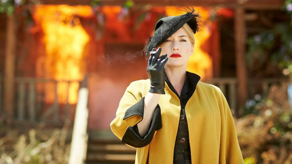 A woman in a yellow coat smoking a cigarette in front of a burning house