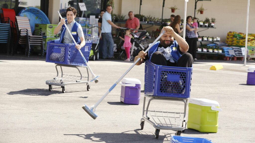 Two men, sitting in shopping carts, racing in a parking lot