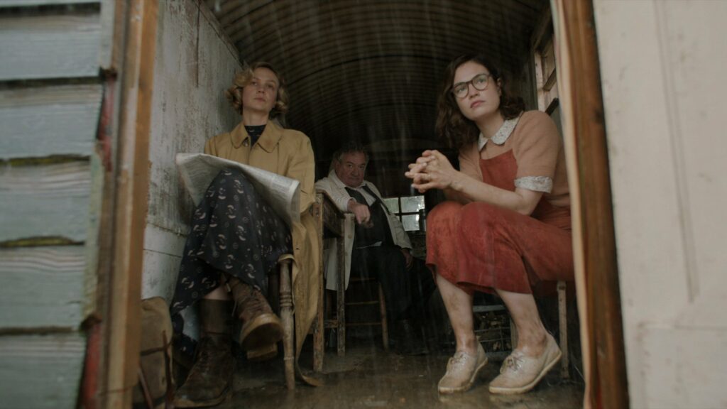 Two women sitting in the dorrway of a house. A man is sitting just within view behind them