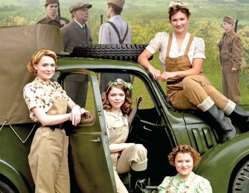 Four women, in 1940s attire, standing beside a military truck