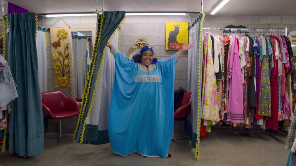 A black woman standing in the doorway of a changing room in a clothing store looking jubilant 