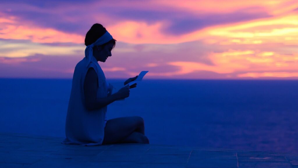 a woman sitting on a beach at sunset reading a letter