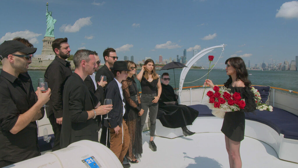 A woman standing on the deck of a yacht, holding an armful of red roses. There is a crowd of people standing on the deck with her.