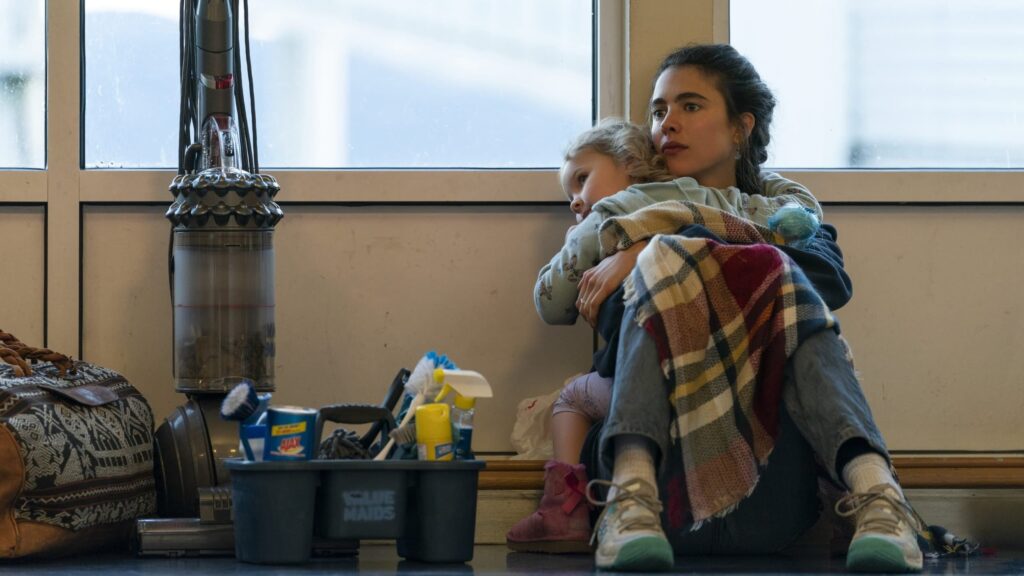 A young woman sitting on the floor of a ferry station, cradling her sleeping toddler